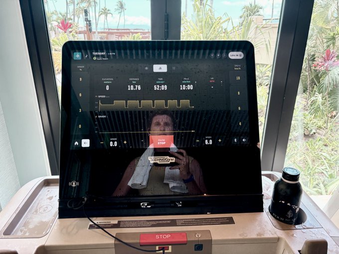 A person takes a mirror selfie on a Technogym treadmill displaying workout stats including elevation 0, distance 10.76, time 52:09, and pace 10:00, with a water bottle beside the console.