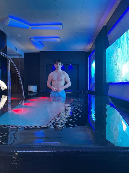 A shirtless man stands waist-deep in a modern, blue-lit indoor pool or spa, surrounded by ambient lighting and tiled floors. The image appears to be a playful "gm" (good morning) post, with the man's calm, centered pose serving as a morning greeting to fellow crypto enthusiasts.