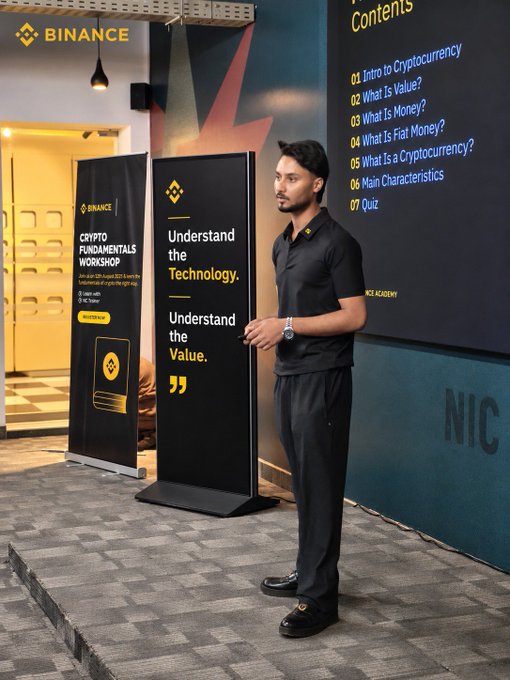A man stands on a stage during a "Binance Crypto Fundamentals Workshop," with banners reading "Understand the Technology. Understand the Value." and a slide listing cryptocurrency topics.