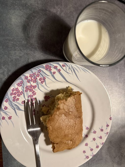 A top-down photo of a pastry or empanada-like baked good on a floral-patterned plate with a fork, alongside a glass of milk on a dark gray surface.