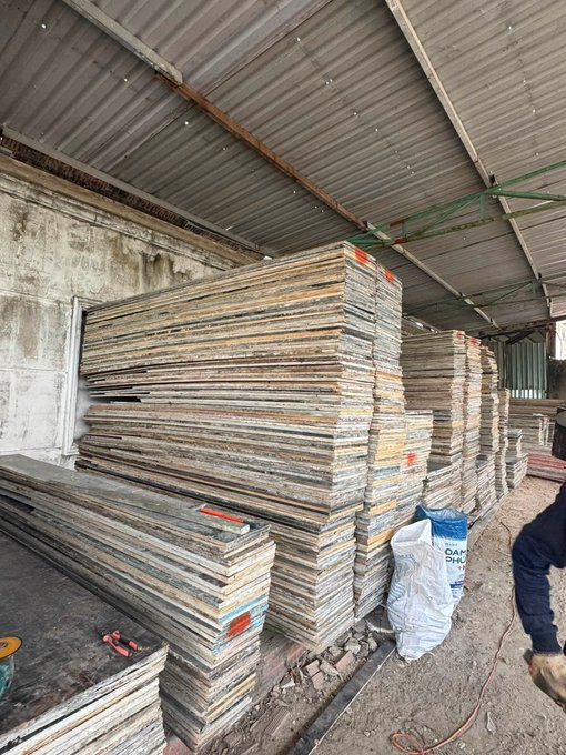 Stacks of weathered construction materials—likely formwork panels or scaffolding boards—piled high under a corrugated metal shed roof, with a partially visible bag labeled "DAM PHR..." sitting nearby. The image contrasts the poster's "grind" in replying online (Web2/Web3 hustle) with an actual physical labor scene of stacked, worn materials waiting to be used or reclaimed.