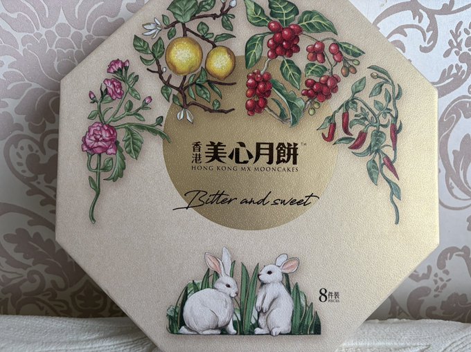 An octagonal Hong Kong MX Mooncakes box (8-piece) decorated with illustrations of fruits, flowers, chili peppers, and two rabbits, with the tagline "Bitter and sweet." The post celebrates receiving these Mid-Autumn Festival mooncakes as a gift from Ave and Yoda, symbolizing gratitude and the recognition of quality products by the market.