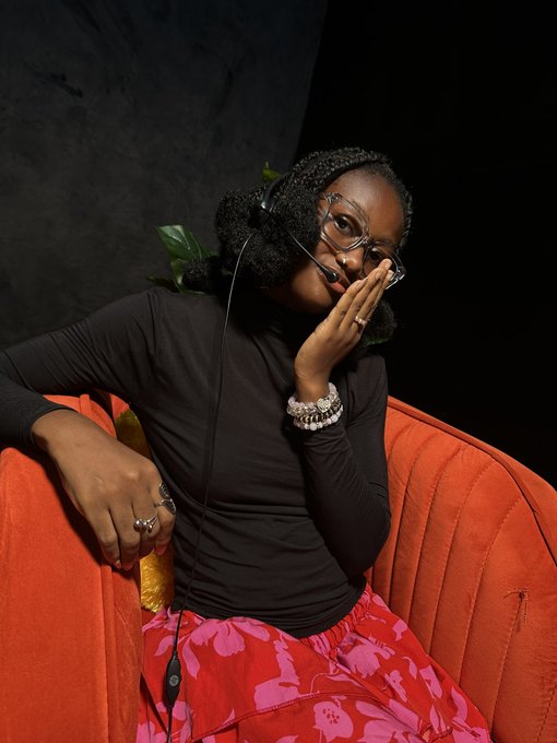 A young woman wearing glasses, headphones, and a black long-sleeve shirt sits on a red couch with a green leaf, appearing to blow a kiss while wearing a red patterned skirt and multiple bracelets.