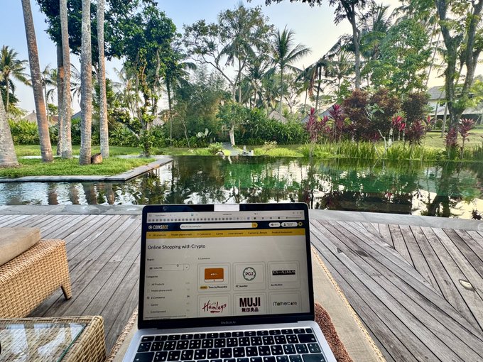 A laptop displays the Coinsbee website on a wooden deck overlooking a tranquil tropical pond, with the page featuring brand logos like MUJI and Metro as the user researches Velo Protocol. The serene outdoor setting contrasts with the digital investigation of the crypto-gift card platform mentioned in the post.