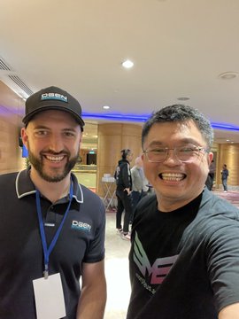 Two smiling men pose for a selfie indoors; one wears a black 'DIGIBOT' cap and lanyard, while the other wears a black t-shirt with 'WE' visible.