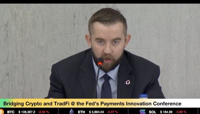 A man in a dark suit speaks at a microphone during the "Bridging Crypto and TradFi @ the Fed's Payments Innovation Conference," with crypto ticker prices displayed below showing BTC at $108,387.2 (-2.53%), ETH at $3,864.04 (-4.37%), and SOL at $184.99 (-3.89%).