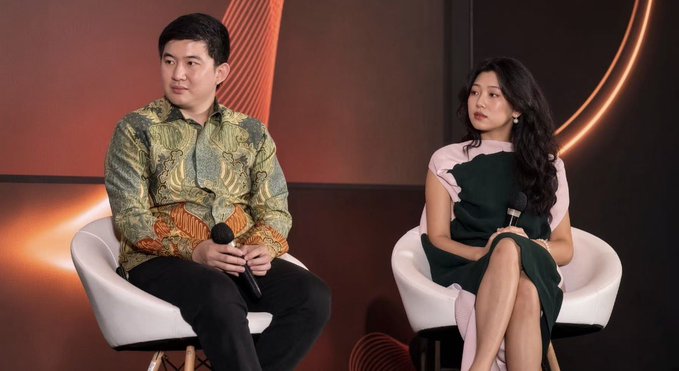 Two people seated on white chairs against an orange backdrop with curved light accents; a man in a batik shirt holding a microphone and a woman in a green and pink dress.