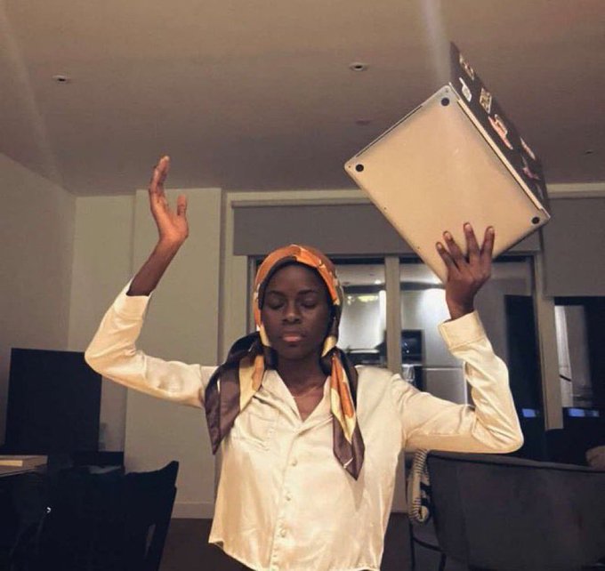 A young woman wearing a headscarf and cream-colored shirt poses indoors, tossing a laptop into the air while holding a phone in her other hand.