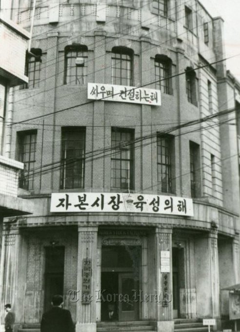 The image features an old building with Asian writing, possibly indicating that it was once part of a Korean business or establishment. The building has two signs above its entrance, one reading "Korea Herald" and another saying "I'VE OPENED A NEW RWA CHANNEL". There is also a person walking in front of the building, adding to the atmosphere of an old-fashioned setting.
