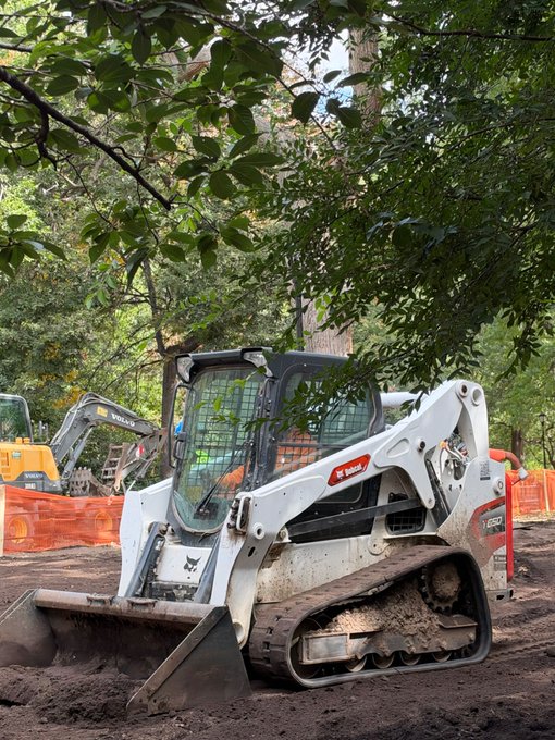 A white Bobcat skid-steer loader parked on a dirt construction site, with another yellow excavator visible in the background among trees.