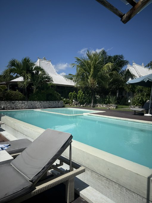 A serene resort pool area with lounge chairs, palm trees, and white-roofed buildings under a clear blue sky, capturing a relaxing tropical getaway.