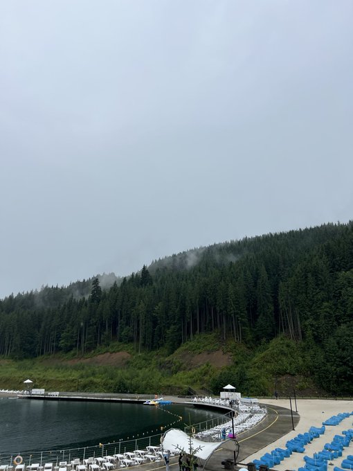 A misty, overcast mountain landscape overlooks a lakeside resort area with rows of blue lounge chairs arranged along the shore.