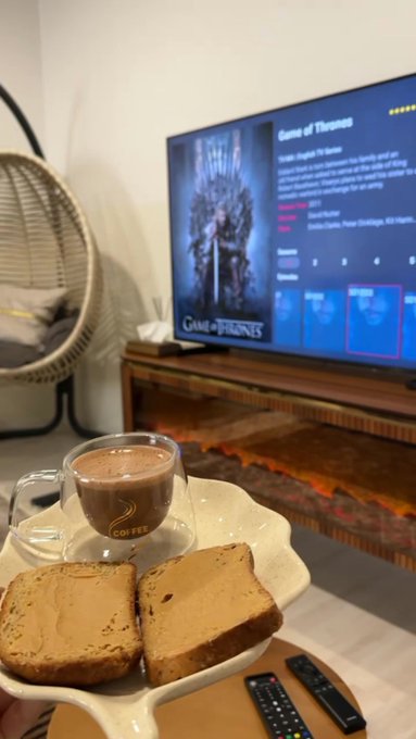 A cozy indoor scene showing a cup of coffee and two slices of toast on a white plate in the foreground, with a TV displaying the Game of Thrones menu screen in the background, next to a hanging egg chair and an electric fireplace.