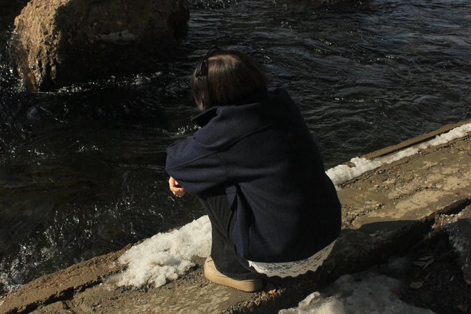 A person with short dark hair, wearing a navy coat and sunglasses perched on their head, crouches by the edge of a cold shoreline with patches of melting snow, gazing out at the dark rippling water. The contemplative, quiet scene reflects the reflective mindset behind the post's morning call to "win today."