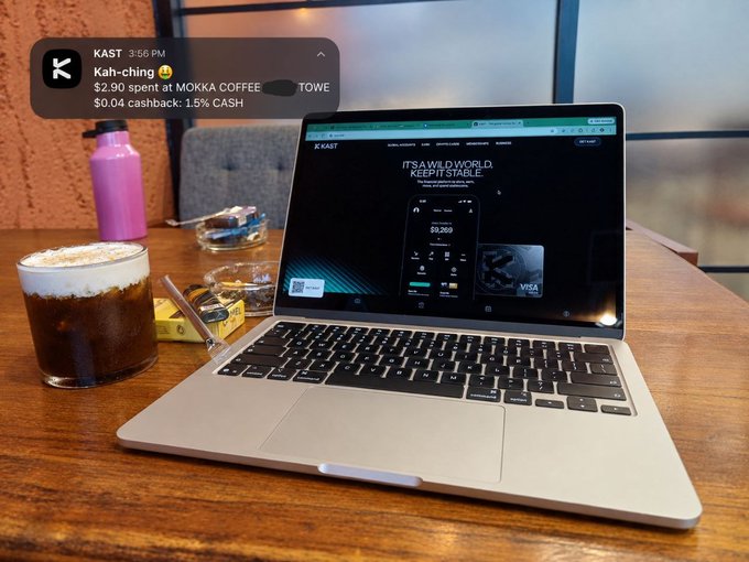 A silver laptop on a wooden table displays the KAST website with the text 'IT'S A WILD WORLD, KEEP IT STABLE.' alongside a coffee drink, a pink water bottle, and a KAST notification overlay showing 'Kah-ching $2.90 spent at MOKKA COFFEE'.