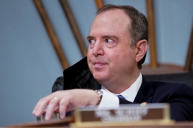 A man in a suit looking to the side during what appears to be a government hearing, with a nameplate partially visible on the desk.