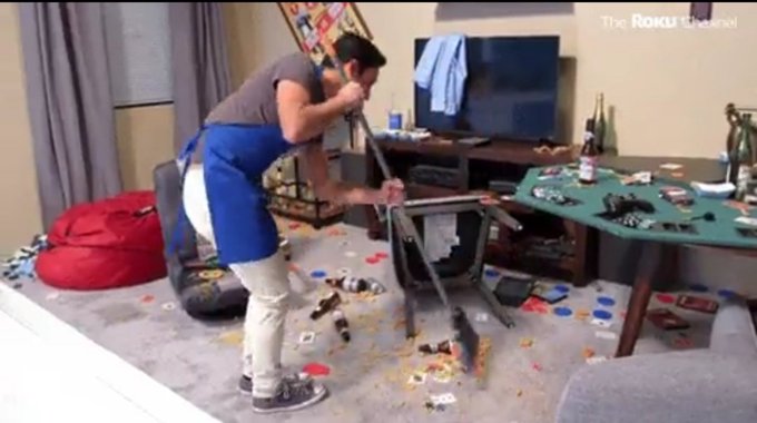 A man in a blue apron stands amid a heavily trashed room with debris scattered across the floor, bottles and items strewn around a table and bean bag.