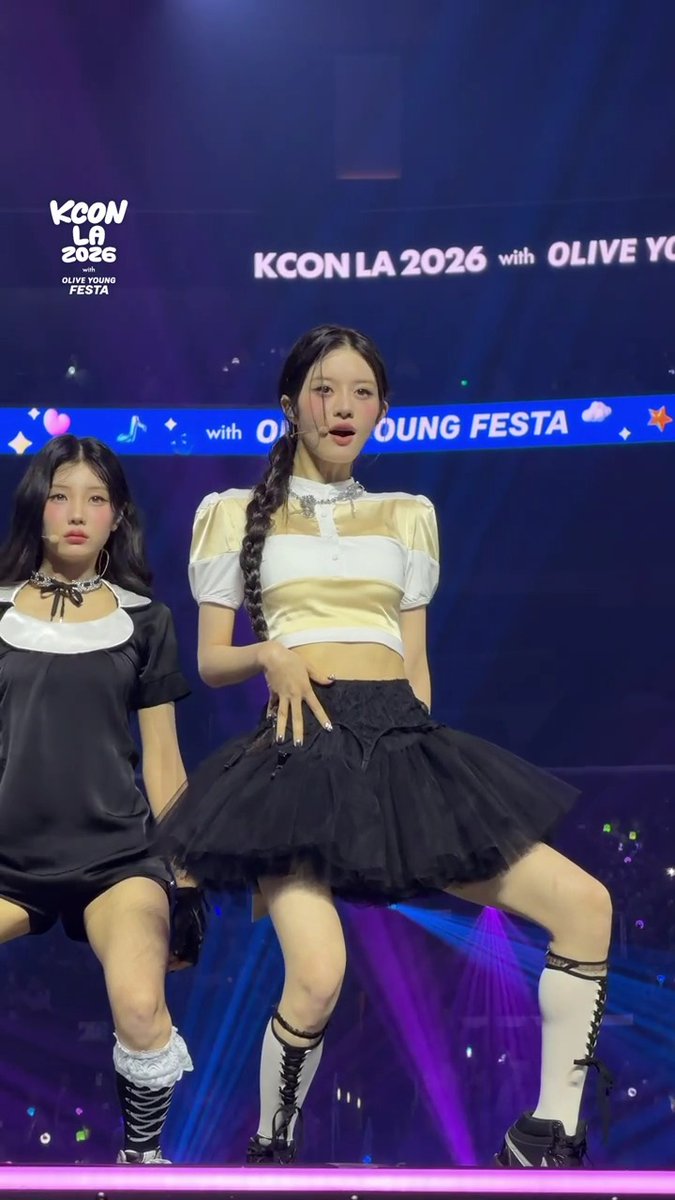 Two performers on stage at KCON LA 2026 with OLIVE YOUNG FESTA; one wears a yellow and white crop top with a black tutu and white lace-up boots, while the other is in a black dress.