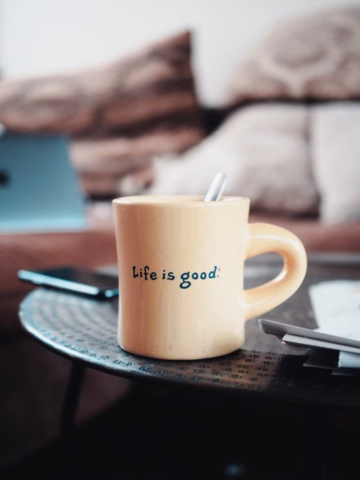 A yellow ceramic mug reading 'Life is good.' sits on a dark round table with a spoon inside, in front of a blurred living room setting with a laptop and couch.