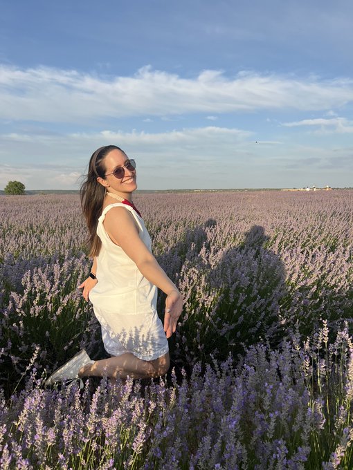 A woman in a white dress and sunglasses smiling in a vast lavender field under a blue sky.