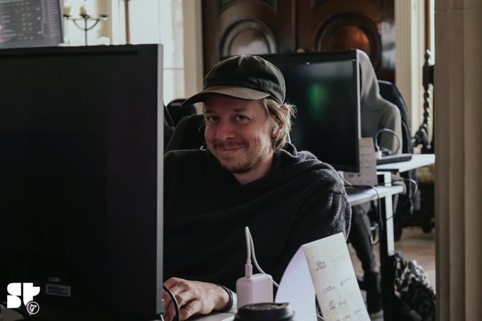A man wearing a black cap and sweater sits at a computer monitor in an office setting, smiling toward the camera with a paper coffee cup and sheet music nearby; an "ST" logo appears in the corner.