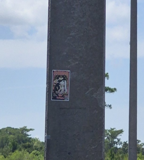 A small poster or sticker is affixed to a dark metal pillar against a cloudy sky, with green trees in the background. The image relates to the post by symbolizing "the city takeover," as the sticker represents the grassroots placement of SPX6900 promotional materials in public spaces.