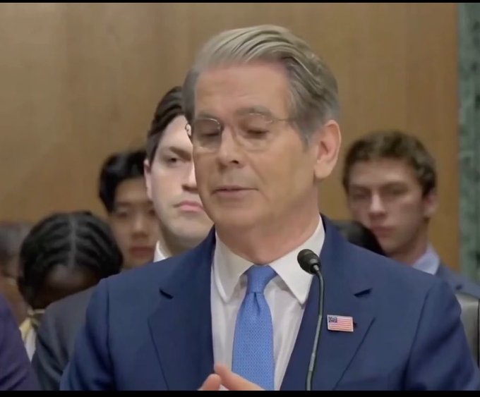 A man in a blue suit with an American flag pin speaks at a microphone, likely during a Senate hearing, with other individuals seated behind him.
