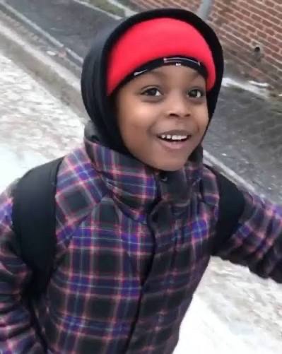 A smiling young boy wearing a red headband and a plaid jacket with backpack straps outdoors in a snowy setting.