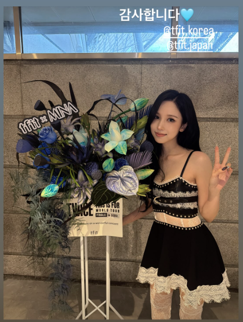 A woman in a black dress poses with a peace sign next to a floral display featuring a "tfit × MINA" sign at what appears to be a TWICE concert, with text on the image reading "감사합니다💙" (Thank you) and tagging @tfit_korea and @tfit_japan, celebrating the TFIT x MINA collaboration event.