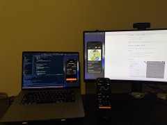 The image shows two computer screens side by side on a desk. One screen displays a webpage with a phone attached to it, while the other screen has a desktop computer setup with a laptop and a monitor. The desktop computer also features a keyboard and mouse. A remote control is placed in front of one of the screens.