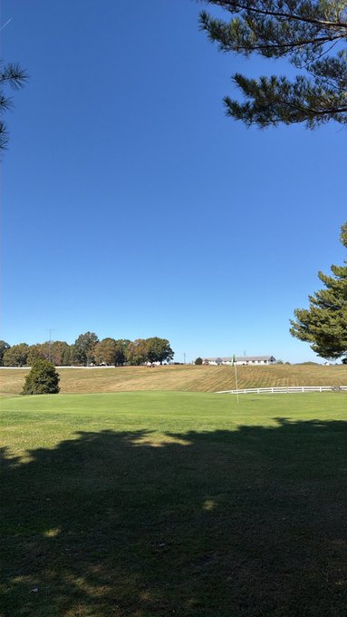A scenic outdoor view of a golf course green with a flag, surrounded by rolling hills, trees, and a clear blue sky with pine branches visible in the upper right corner.
