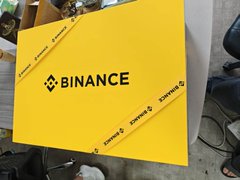 The image features a yellow box with "BNB" written on it, which could be related to Bitcoin or another cryptocurrency. The box has a ribbon tied around its neck, indicating that it might have been recently opened or is being presented for sale. There are two people in the background of the image - one person sitting at a desk and another standing near the box.
