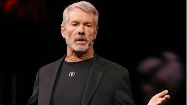 A photograph of an older man with grey hair and beard wearing a black blazer over a dark shirt with a small logo, appearing to speak at an event.