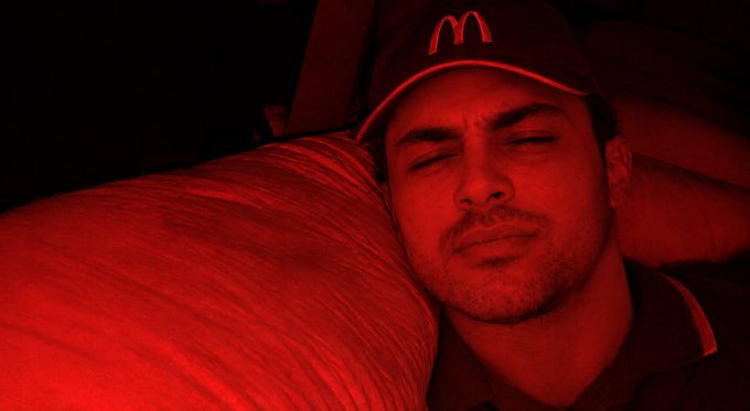 A man wearing a McDonald's cap appears to be sleeping on an airplane, with the cabin illuminated by red lighting.