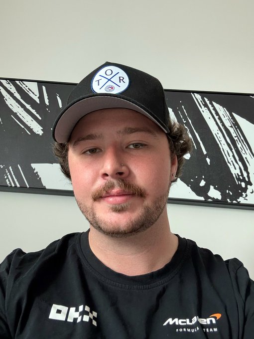A young man wearing a McLaren Formula Team shirt and a black cap with a "TQR" logo takes a selfie with a subtle smile. The "GM" (good morning) greeting paired with the image suggests he's sharing a casual, upbeat start to his day, likely as a Formula 1 fan showing off his McLaren apparel.