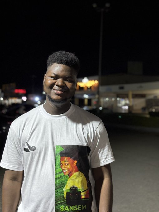 A smiling young man poses for a photo at night in what appears to be a parking lot, wearing a white t-shirt featuring an image of crypto influencer Ansem with the text "$ANSEM" printed below it. The image directly reflects the post's caption about being out late "in the trenches" onboarding people and sharing $ANSEM tips, with the shirt serving as a visual shoutout to the crypto community.