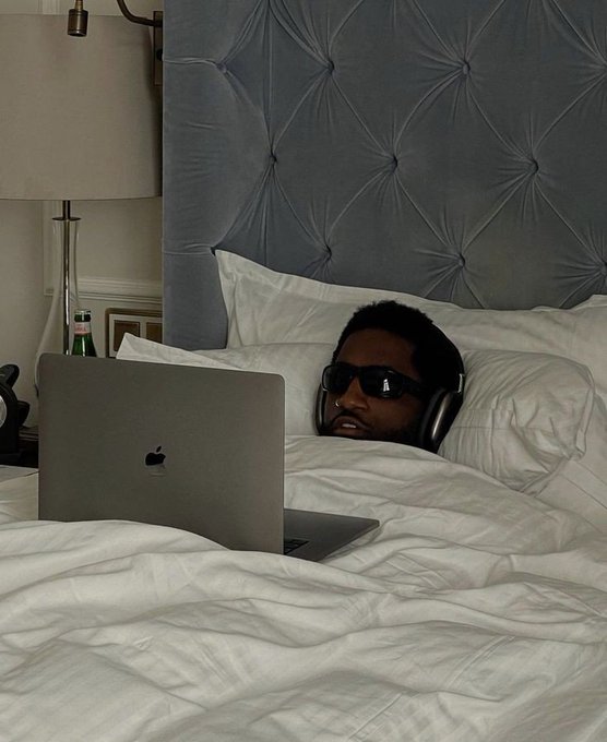 A man wearing dark sunglasses lies in a luxurious bed with a tufted grey headboard, using a silver Apple MacBook laptop.