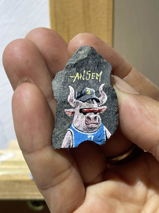 Fingers hold a small piece of slate rock with a hand-painted cartoon of a pig wearing a blue tank top, sunglasses, and a backwards cap, with the word 'ANSEM' written in yellow above it.