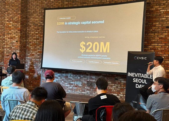 The image shows a group of people gathered around a large projector screen, with a speaker presenting information about strategic capital secured. The numbers displayed on the screen are $20m. A woman standing next to the man at the podium appears to be talking about the project's execution and expansion plans for the ecosystem. There is also an advertisement visible in the image that reads "xeffy just dropped and the numbers are already talking $20m in strategic capital secured". Additionally, a crowd of people can be seen sitting down behind the man giving his presentation, along with two chairs positioned near them.
