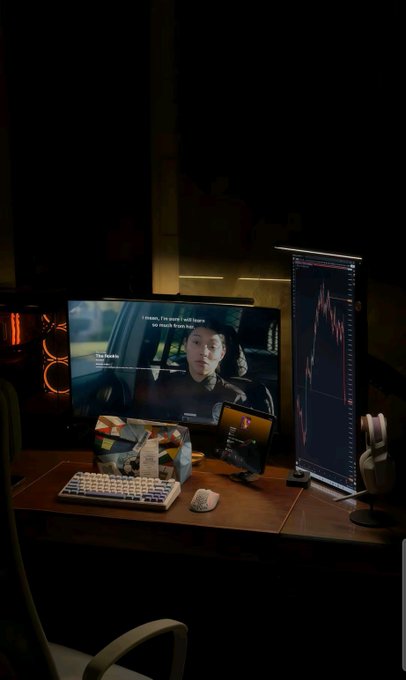 A dark, dimly lit gaming or workstation setup featuring a wide monitor displaying a video game scene with subtitle text, alongside a vertical monitor showing a financial chart, with a keyboard, mouse, and tablet on the wooden surface.