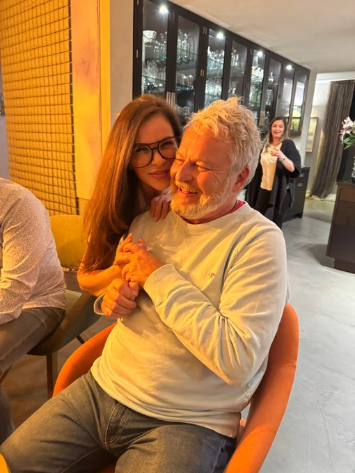 An older man with white hair and a beard in a white sweater laughs as a woman with long brown hair and glasses affectionately holds his hands indoors, with another woman visible in the background.