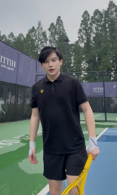The image features a young man standing on a tennis court, holding a yellow tennis racket. He appears to be posing for a photo or participating in an event related to tennis. The background of the image shows trees, suggesting that the tennis court is located near a park or natural area. There are also words visible in the image, such as "Binance", "binance真的很时尚", and "@0xWatell @sisibinance @pear_bnb".