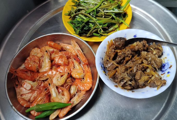 A metal tray holds three dishes: a bowl of boiled shrimp with green chilies, a yellow plate of cooked greens, and a bowl of braised meat with ginger.