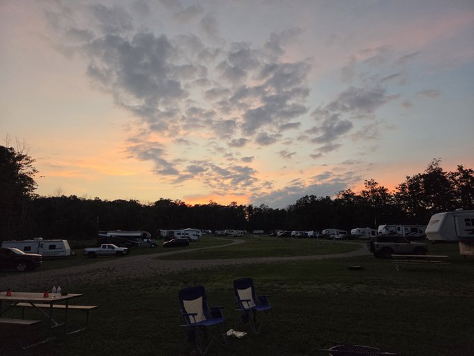 A scenic view of an RV campground at sunset, featuring a colorful sky with pink and orange hues above scattered clouds, with campers, vehicles, picnic tables, and empty chairs on the grassy field.