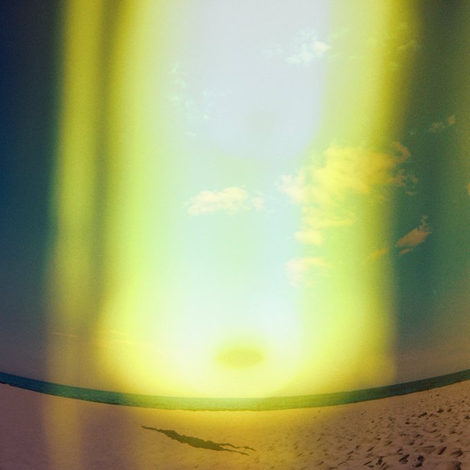 A dreamlike beach scene with a bright yellow and green glowing light, footprints in the sand, and clouds visible in the sky.