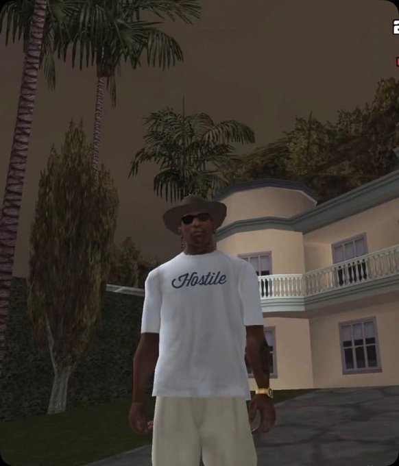 A screenshot from a video game showing a male character wearing a white t-shirt with "Hostile" written on it, a hat, and sunglasses, standing in front of a large house with palm trees.