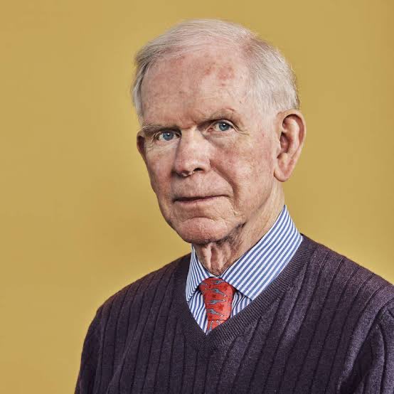 The image shows an older man with white hair wearing a purple sweater over a striped shirt and red tie, posing against a yellow background. The caption doesn't directly relate to the post about Bitcoin price predictions, as the image appears to be an unrelated portrait rather than anything containing visible text, numbers, or tickers.