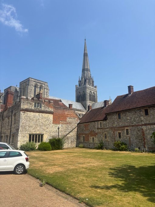 A historic stone cathedral with a tall pointed spire stands behind old brick and stone structures under a clear blue sky, with a white car parked on a gravel driveway in the foreground.
