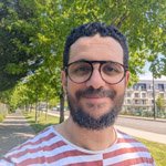 A man with glasses and a beard smiles while taking a selfie outdoors, wearing a red and white striped shirt.