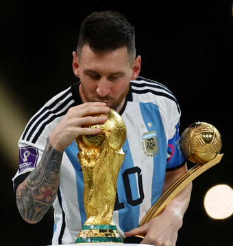 Lionel Messi, wearing Argentina's #10 jersey, kisses the FIFA World Cup trophy after a victory, holding the golden trophy with the iconic two-hand design.